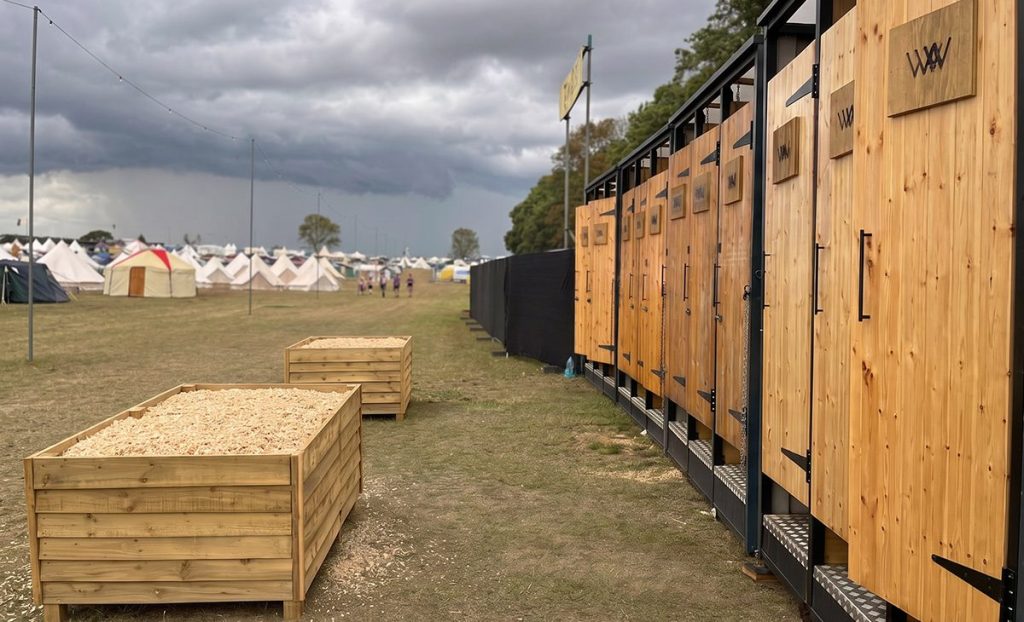 Row of wooden restroom facilities at an outdoor event, with tents visible in the background and stormy skies overhead.