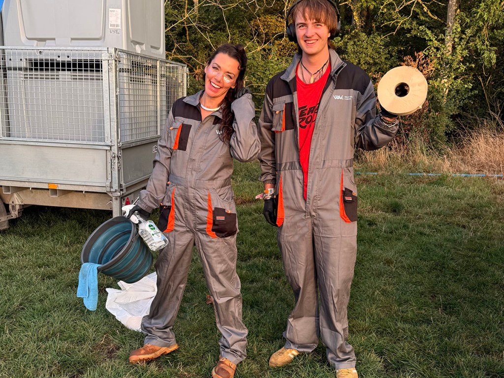 Two individuals wearing gray and orange work overalls stand outdoors, smiling. One holds a blue container and a milk carton, while the other holds a roll of material. A trailer is visible in the background.