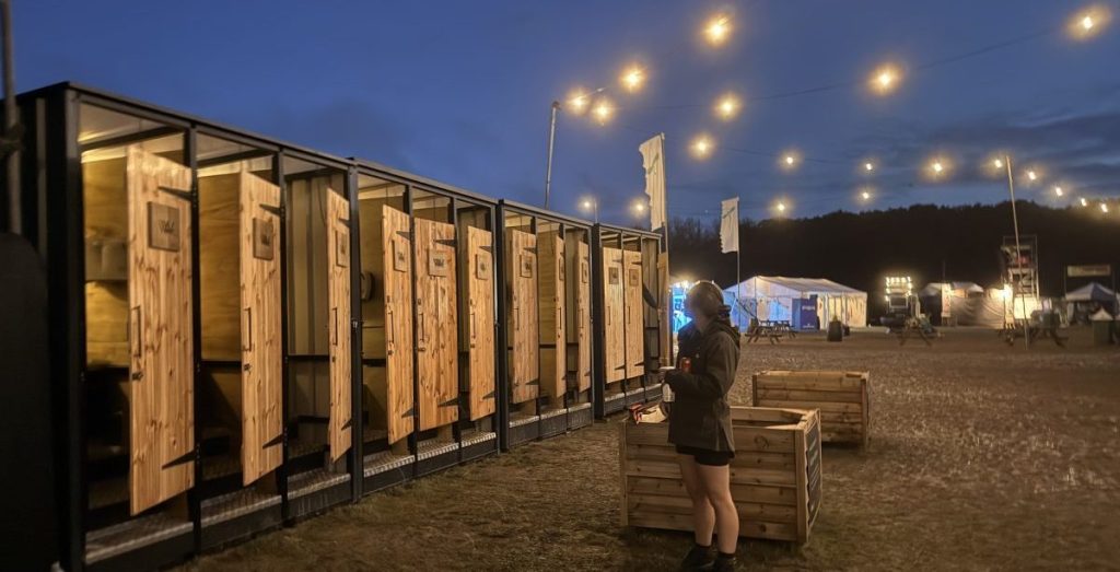 A person standing in front of a row of wooden restroom units at dusk, with string lights illuminating the area and festival tents in the background.
