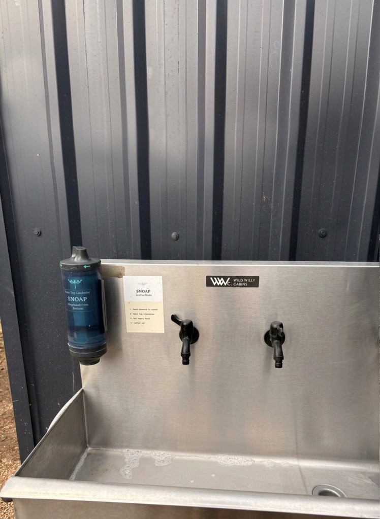 Outdoor handwashing station with a soap dispenser and two taps, mounted on a metallic surface.