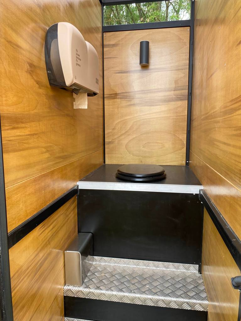 Interior of a portable restroom featuring wooden paneling, a toilet with a black seat, and a paper towel dispenser mounted on the wall.